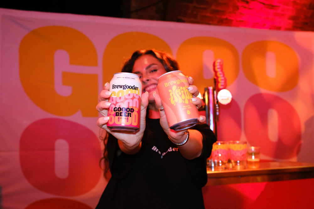 Person holding two cans of Good Beer in front of a colorful background with 'Good Beer' branding.
