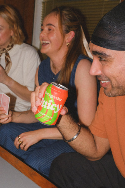 A group of three people sitting together laughing, one holding a can of Brewgooder Juicy, in a casual setting. Photo taken using a flash and edited in 35mm film style