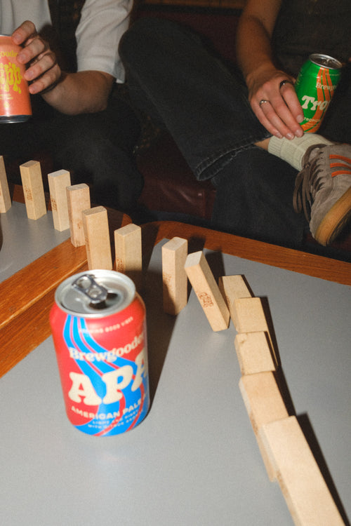 an out-of-focus can of Brewgooder American Pale taken using a flash and edited in a 35mm film style, there are Jenga blocks set up as dominos falling in a line behind the beer